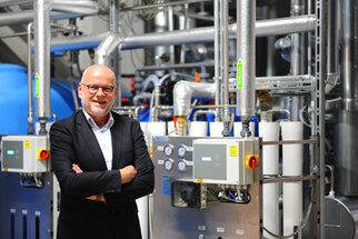 A man in a suit stands smiling in front of an industrial water treatment facility, surrounded by pipes and control equipment.