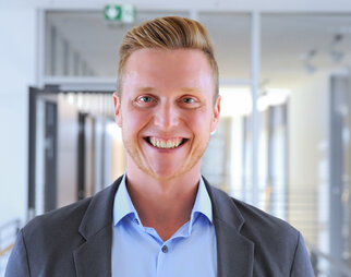 A smiling man with blonde hair is wearing a gray suit and a light blue shirt. He is standing in a modern interior.