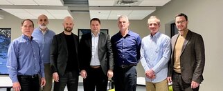 A group of seven men stands in a modern room, all dressed in formal attire, wearing a variety of shirts and suits.