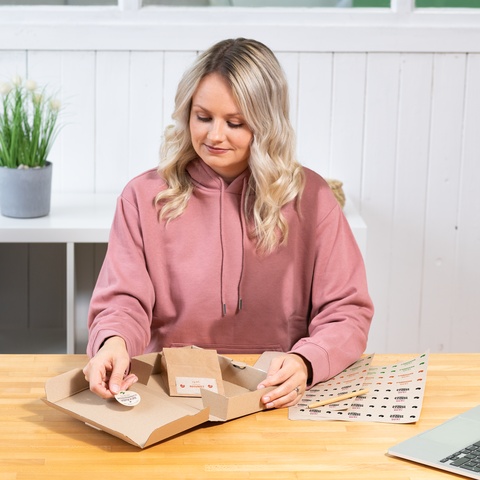 A person with blonde, curly hair is sitting at a table, working on a package while looking at stickers.