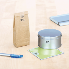 Ein silberner Metallbehälter, ein brauner Papiersack mit Logo, ein grünes Blatt und ein blauer Stift auf einem hellen Tisch.