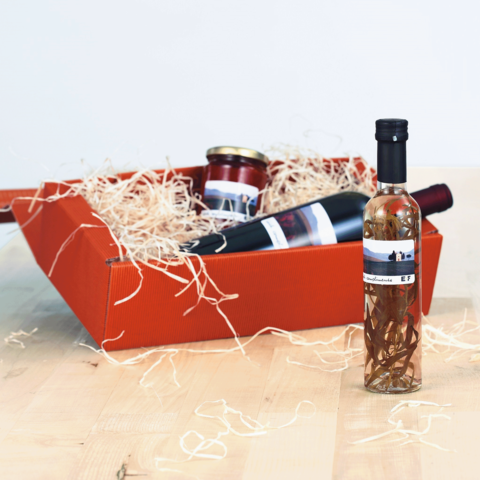 Ein roter Geschenkkorb mit einer Flasche Likör, einem Glas Marmelade und einer Weinflasche, umgeben von Holzwolle.