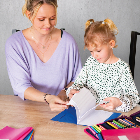 A woman and a little girl are sitting at a table, flipping through a notebook. Colorful pens are scattered across the table.