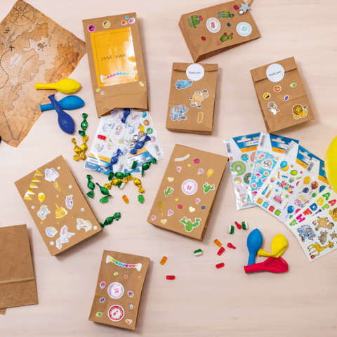 Bastelmaterialien auf einem Tisch, darunter Papiertüten, Aufkleber, Scheren, Ballons und bunte Dekorationselemente.