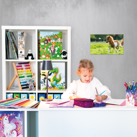 Ein Mädchen sitzt an einem Schreibtisch, schreibt in ein Heft und hat bunte Papiere und Stifte um sich herum. Im Hintergrund sind Regale mit Spielzeug.