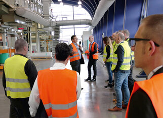 A group of people in safety vests is listening to a speaker in an industrial setting.
