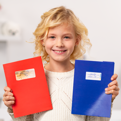 A smiling child holds a red notebook and a blue notebook in their hands, both covered in stickers.