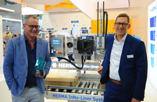 Two men are standing next to a HERMA InNo-Liner system, a machine for labelling, while one of them is holding a prize.