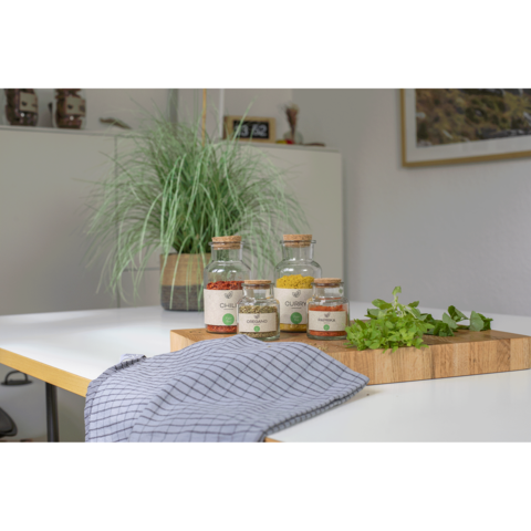 Glasbehälter mit Gewürzen und frischen Kräutern auf einem Holzbrett, daneben ein gestreifter Küchentuch und eine Pflanze im Hintergrund.