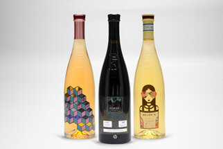 Three bottles of wine stand side by side: one pink, one black, and one yellow, each with a unique, artistic label.