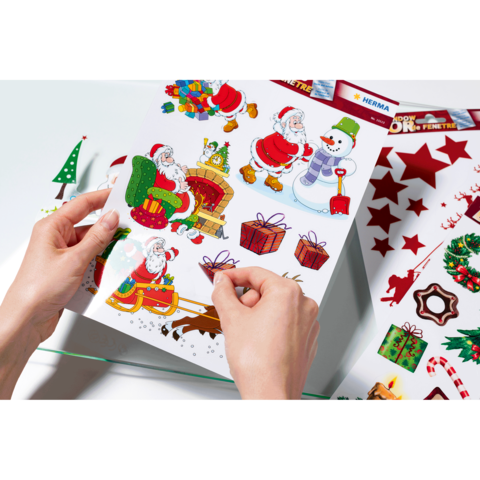 Hände halten ein Blatt mit bunten Weihnachtsstickern, darunter Weihnachtsmänner, Geschenke, einen Schneemann und Weihnachtsdekorationen.
