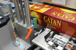 A packaging machine processing a box of the game 'Catan Plus,' with various mechanical components in the foreground.