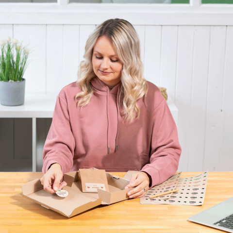 Eine Frau mit blonden, lockigen Haaren sitzt an einem Tisch und öffnet eine Kartonverpackung. Auf dem Tisch liegen Aufkleber und eine Pflanze im Hintergrund.