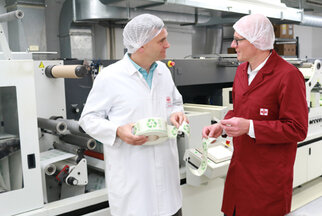 Two people in protective gear are standing in a production environment discussing eco-friendly packaging labels.