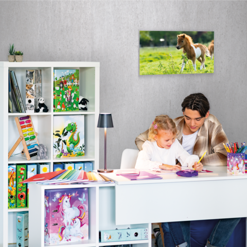Ein Kind und ein Erwachsener sitzen an einem Schreibtisch und malen. Im Hintergrund steht ein Regal mit Spielzeug und Büchern. An der Wand hängt ein Bild eines Pferdes.
