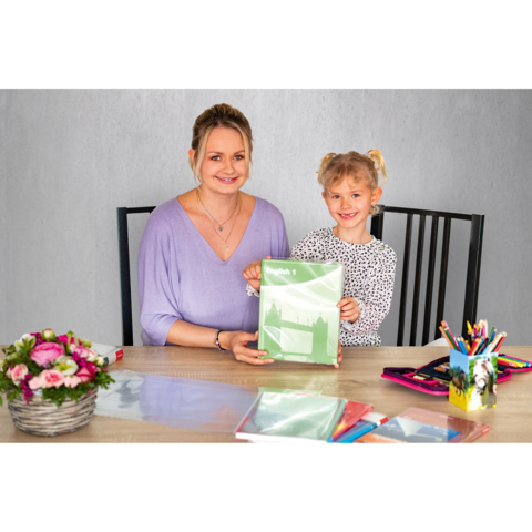 Eine Frau und ein Mädchen sitzen an einem Tisch, beide lächeln und halten ein grünes Heft. Auf dem Tisch liegen Schreibmaterialien und Blumen.