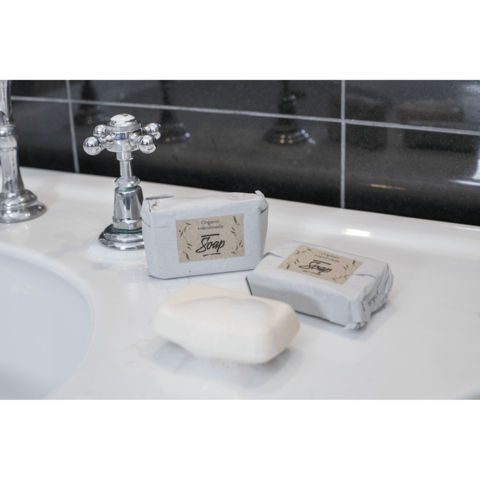 Zwei Seifenstücke, eines in grauem Papier verpackt, liegen auf einem Waschbecken neben einem weißen Seifenstück und einem Wasserhahn.
