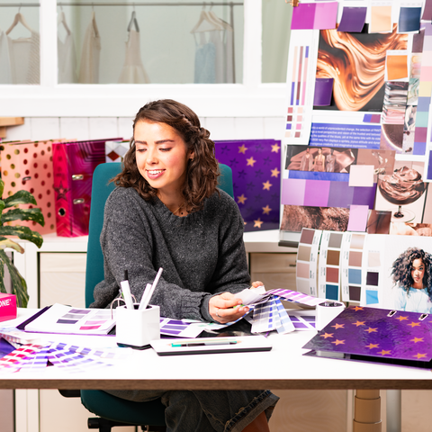 A woman with curly hair sits at a table, surrounded by color swatches and design materials, as she works on a project.