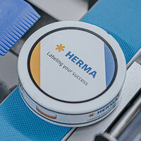 Round container with the print 'HERMA' and the slogan 'Labelling your success,' surrounded by blue and white elements.