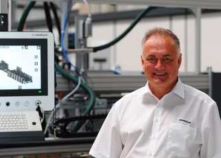 A man in a white shirt stands smiling in front of a screen displaying technical information, in an industrial setting.
