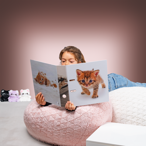 A child is sitting in a large, round bean bag chair, reading a folder with a picture of a kitten on the front.