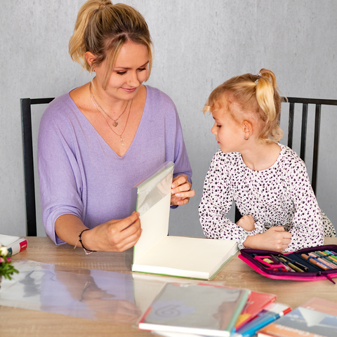 Eine Frau und ein kleines Mädchen sitzen an einem Tisch, während sie gemeinsam an einem kreativen Projekt arbeiten. Bunte Stifte und ein Blumenarrangement sind sichtbar.