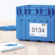 Blaue Kunststoffbox mit einer Etikette, die die Nummer 0134 und zusätzliche Informationen enthält, auf einem Tisch neben einem Notizblock und einem Stift.