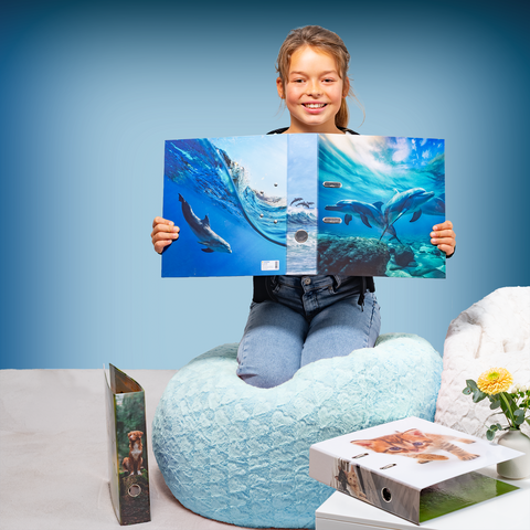 A girl is sitting on a bean bag holding a book with pictures of dolphins. Next to her, there are more books on a table.