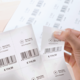 Ein Hand hält ein Etikett mit Informationen zu einem Kleidungsstück, einschließlich Preis, Material und Größe, während weitere Etiketten im Hintergrund sichtbar sind.