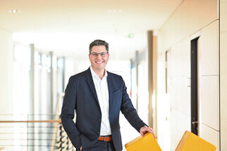 A man in a suit stands in a modern hallway, smiling as he holds onto the back of a yellow chair.