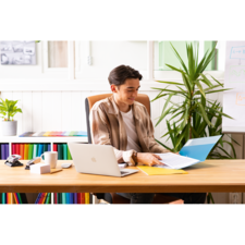 Ein junger Mann sitzt an einem Schreibtisch, arbeitet mit einem Laptop und blättert durch Dokumente in einer Ordnermappe.