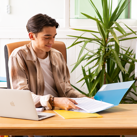 Ein junger Mann sitzt an einem Schreibtisch, arbeitet mit einem Laptop und blättert durch Dokumente in einer Ordnermappe.