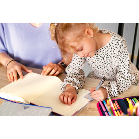 Ein kleines Mädchen schreibt in ein offenes Notizbuch, während eine erwachsene Person daneben sitzt und zuschaut. Bunte Stifte liegen auf dem Tisch.