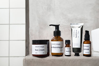A selection of natural cosmetic products, including face cream, cleansing gel, tooth oil, hand cream, and serum, displayed on a gray surface.