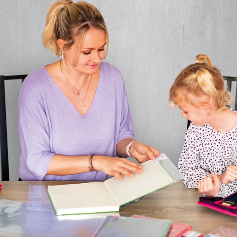 Eine Frau und ein Mädchen sitzen an einem Tisch und betrachten gemeinsam ein Buch. Auf dem Tisch liegen weitere Bücher und Bastelmaterialien.