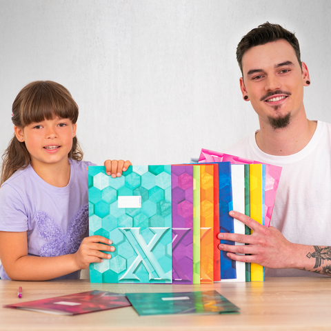 Ein Kind und ein Mann halten bunte, geometrisch gestaltete Ordner und präsentieren sie lächelnd vor einem Tisch.