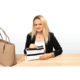 Eine Frau mit langen blonden Haaren sitzt an einem Tisch und hält eine durchsichtige Tasche in der Hand. Neben ihr liegt eine große Handtasche.