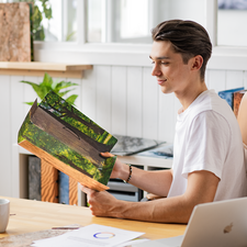 Ein junger Mann hält ein Buch mit einem Waldmotiv in der Hand, während er an einem Tisch sitzt. Im Hintergrund sind Pflanzen und Möbel sichtbar.