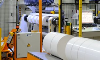 Industrial machines for processing roll material, with a control panel in the background and safety fencing.