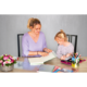Eine Frau und ein Mädchen sitzen an einem Tisch und betrachten gemeinsam ein Buch. Auf dem Tisch liegen verschiedene Schreibmaterialien und Blumen.