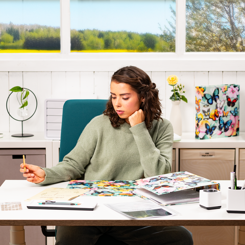 A young woman sits at a desk, surrounded by colorful materials and notes, while thoughtfully holding a pen.