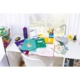 Ein Schreibtisch mit bunten Ordnern, Schreibmaterialien, einem Notizbuch und einer Lampe. Im Hintergrund sind Fenster und eine kleine Blume sichtbar.