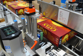A production line with boxes of the game 'Catan Plus' being processed by machines.