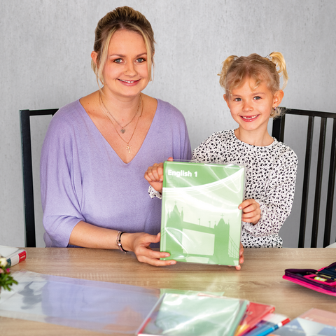 Eine Frau und ein Mädchen sitzen an einem Tisch, das Mädchen hält ein grünes Heft. Auf dem Tisch liegen Schreibwaren und Blumen.