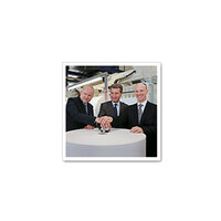 Three men in suits stand around a table, placing their hands on an object in the centre.