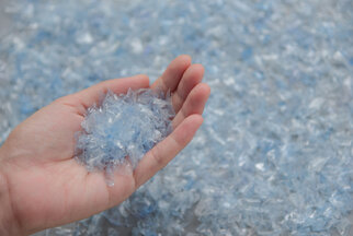 A hand holds a bunch of blue, transparent plastic particles, surrounded by more similar particles on a surface.