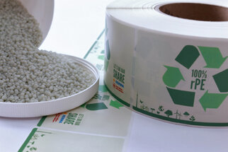 Green plastic pellets are in an open container, next to a roll of eco-friendly labels displaying recycling symbols.