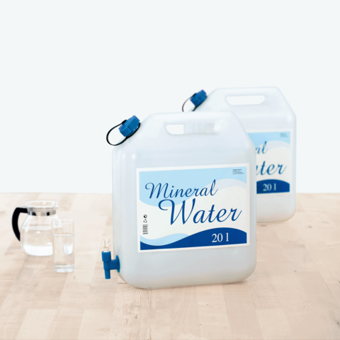 Zwei große Behälter mit Mineralwasser, jeweils 20 Liter, stehen auf einem Tisch. Ein Glas und eine Karaffe sind ebenfalls sichtbar.