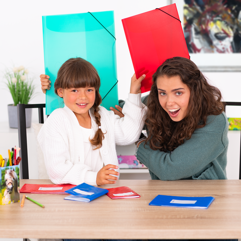 Zwei Mädchen sitzen an einem Tisch, halten bunte Ordner über ihren Köpfen und lächeln. Auf dem Tisch liegen mehrere Hefte in verschiedenen Farben.