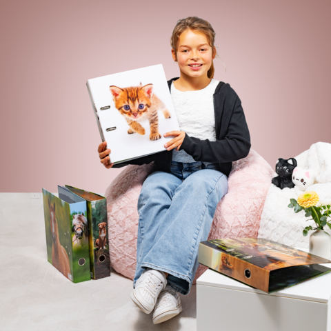 A girl is sitting on a bean bag, holding a binder with a kitten on the cover. Around her are more binders and a plant.